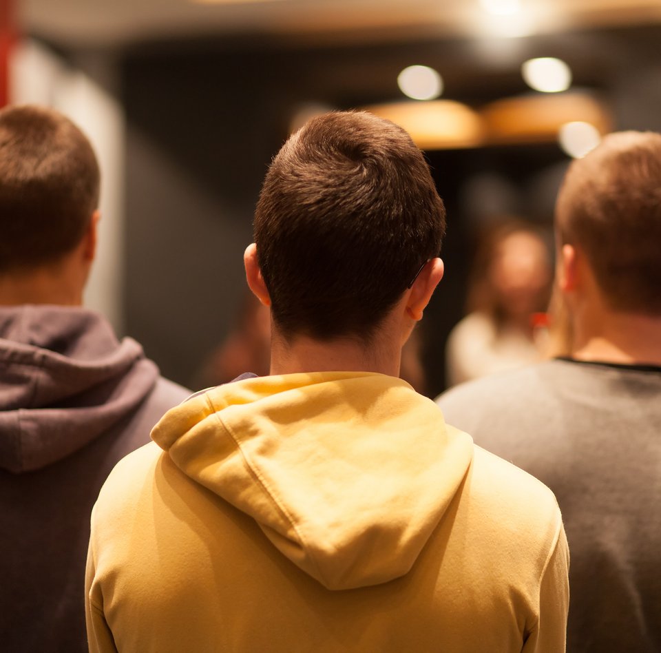 Auf der Farbfotografie sind drei junge Männer zu sehen. Sie sind von hinten fotografiert. Sie befinden sich in einem Ausstellungsraum, der verschwommen im Hintergrund angedeutet ist.