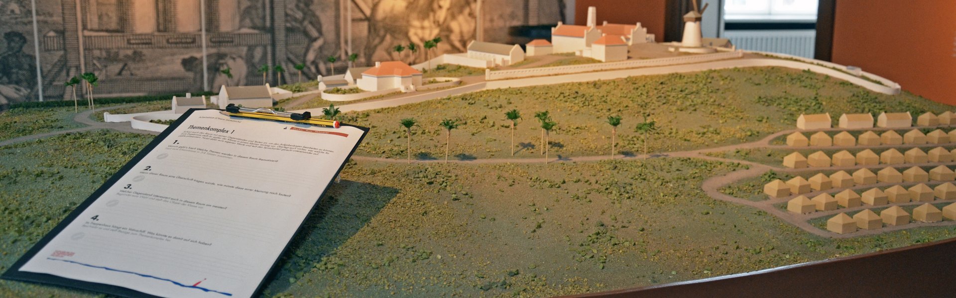 Farvefotografi, der viser et udsnit af udstillingen. I midten af rummet står en model. Den viser en sukkerrørsplantage med marken og mange små og et par store bygninger. På modellen ligger et klembræt. På klembrættet er der fastgjort et stykke papir med spørgsmål og en blyant. I baggrunden af fotografiet ses en tapet. Den viser en sukkermølle, der drives af dyr.