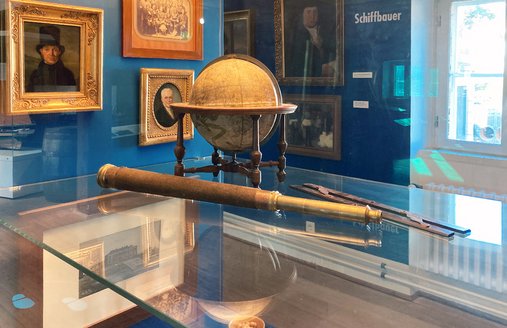 En kikkert og en globus står i en vitrine. På den blå sidevæg hænger billeder af mennesker.