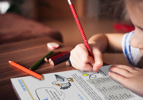 Ein Mädchen hält einen Stift in der Hand und zeichnet damit im Logbuch des Flensburger Schifffahrtsmuseum. Dieses ist aufgeschlagen und zeigt eine farbige Seite mit Texten, Aufgaben und Bildern. Auf dem Tisch liegt nicht nur das Logbuch, sondern auch Buntstifte und ein Radiergummi.