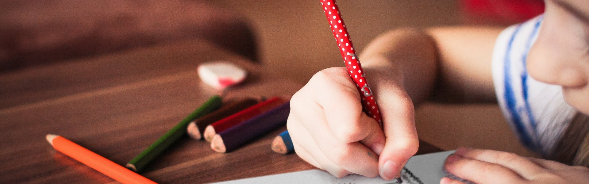 Ein Mädchen hält einen Stift in der Hand und zeichnet damit im Logbuch des Flensburger Schifffahrtsmuseum. Dieses ist aufgeschlagen und zeigt eine farbige Seite mit Texten, Aufgaben und Bildern. Auf dem Tisch liegt nicht nur das Logbuch, sondern auch Buntstifte und ein Radiergummi.