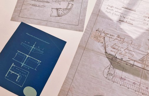 Auf einem weißen Tisch liegen verschiedene Zeichnungen. Sie zeigen: So wir ein Schiff gebaut. Eins der Papiere ist blau und die Zeichnung weiß. Beide anderen Papiere sind weiß.