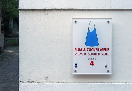 Auf der bunten Fotografie ist eine weiße Gebäudemauer zu sehen. An dieser ist das Stationsschild mit der Nummer 4 der RUM & ZUCKER MEILE befestigt. Auf diesem Schild ist ein großer Zuckerhut als Illustration gedruckt.