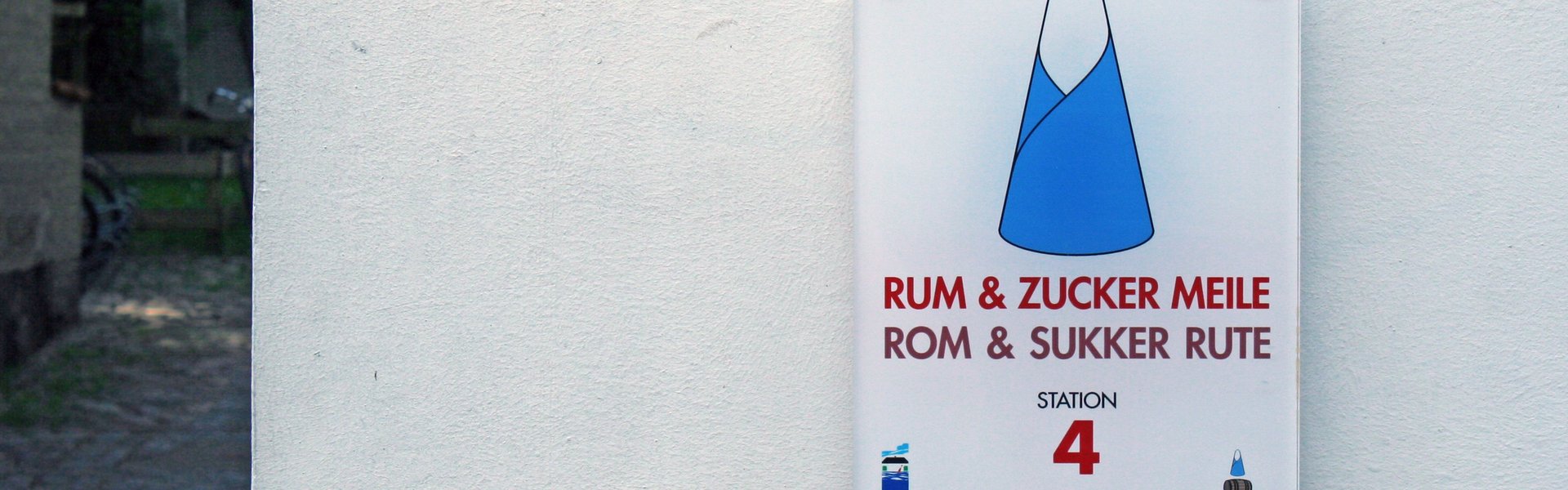 Auf der bunten Fotografie ist eine weiße Gebäudemauer zu sehen. An dieser ist das Stationsschild mit der Nummer 4 der RUM & ZUCKER MEILE befestigt. Auf diesem Schild ist ein großer Zuckerhut als Illustration gedruckt.