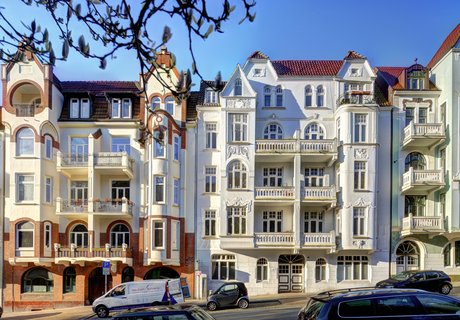 Eine bunte Fotografie. Darauf zu sehen ist eine Häuserreihe. Die Architektur der Häuser ist im Jugendstil. 