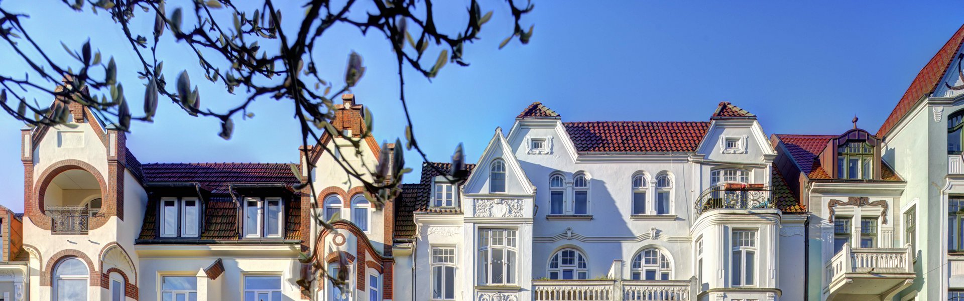 Eine bunte Fotografie. Darauf zu sehen ist eine Häuserreihe. Die Architektur der Häuser ist im Jugendstil. 