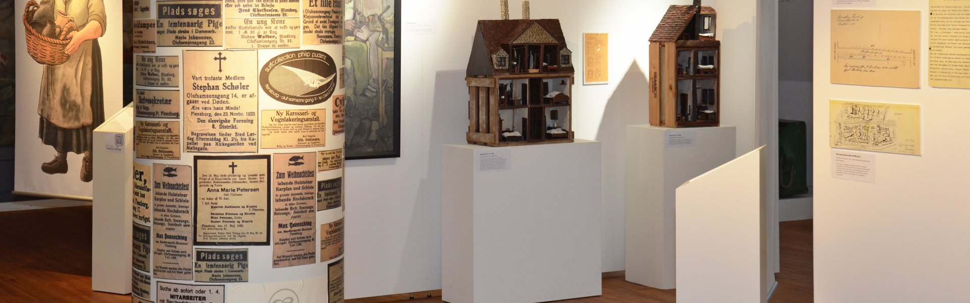 In der Mitte der Ausstellung IDYLLE UND KONFLIKT steht eine Litfaßsäule, die mit verschiedenen alten Zeitungsanzeigen beklebt ist. Alle handeln von dem Oluf-Samson-Gang. Dahinter stehen Modellhäuser zweier Häuser aus dem Gang. Sie stehen vor einer Stellwand. An dieser hängt ein Gemälde, das einen Schuster zeigt. Links im Hintergrund ist ein großes Banner zu sehen. Es zeigt eine mittelalte Frau, die in der Hand einen Korb mit Töpferware trägt. Ihre Kleidung ist schlicht und ein wenig schmutzig.