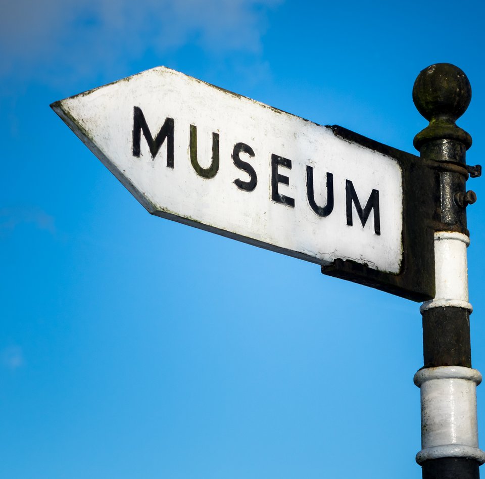 Auf dem bearbeiteten Foto ist das obere Ende eines Wegweisers zu sehen. Der Pfahl ist schwarz-weiß-geringelt. Auf dem Schild, das an dem Pfahl montiert ist, steht in großen Lettern "MUSEUM". Im Hintergrund kann man den blauen Himmel mit ganz zarten Wolken sehen.
