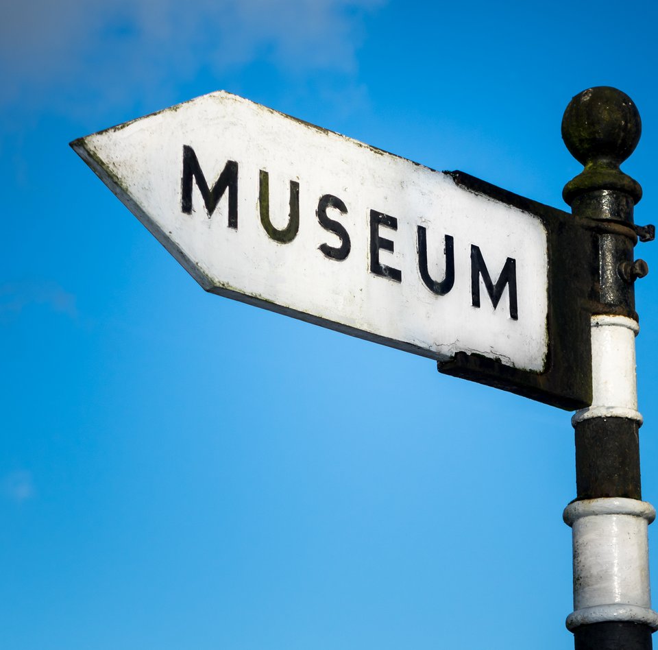 Auf dem bearbeiteten Foto ist das obere Ende eines Wegweisers zu sehen. Der Pfahl ist schwarz-weiß-geringelt. Auf dem Schild, das an dem Pfahl montiert ist, steht in großen Lettern "MUSEUM". Im Hintergrund kann man den blauen Himmel mit ganz zarten Wolken sehen.