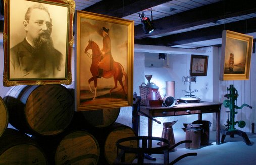 Romtønder er stablet i baggrunden. Foran dem hænger to billeder. Det ene viser en mand med skæg og briller, det andet en mand, der sidder på en hest. To yderligere billeder hænger helt til venstre. Det ene viser et sejlskib på havet, det andet et sejlskib tæt på land. Alle disse billeder har forbindelse til Flensborgs romhistorie. Desuden ses forskellige beholdere og maskiner, der er blevet brugt til aftapning af rom.