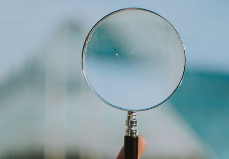 Eine Lupe wird in der Hand gehalten. Von der Hand sieht man nur einen einzigen Finger. Der Hintergrund ist total verschwommen. Man kann lediglich ein weißes Gebäude erahnen.