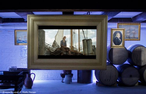 På en stor skærm, der hænger ned fra loftet i romkælderen, vises en film. I scenen ser man en ung mand, der sidder på rælingen af et gammelt sejlskib. Sejlbåden er fortøjet til en pæl. Ved siden af skærmen hænger tre malerier med guldrammer. På dem ser man personer og sejlskibe. Til venstre og højre for den midtplacerede skærm står tønder og andre beholdere til opbevaring af rom.