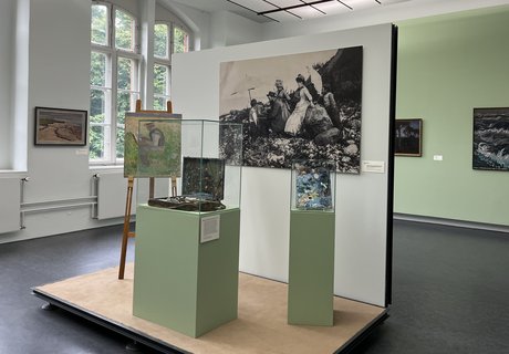 Eine bunte Fotografie. Ein Museumsraum. Im Hintergrund ist auf der linken Seite ein altes großes Fenster. Auf der rechten Seite hängen zwei Gemälde an der Wand. Im Vordergrund ist ein kleines Podest mit Wand. An der Wand hängt eine große Schwarz-weiß-Fotografie eines Mannes der im Freien an einem Bild auf einer Staffelei arbeitet. Um ihn herum stehen Frauen und Kinder in Kleidern, die man um 1900 getragen hat. Auf dem Podest ist eine Staffelei. Darauf das Bild eines Mannes beim Malen. Daneben ist eine Vitrine mit einem alten Malkoffer. Ganz rechts ist eine kleinere Vitrine mit einer alten Malerpalette.  