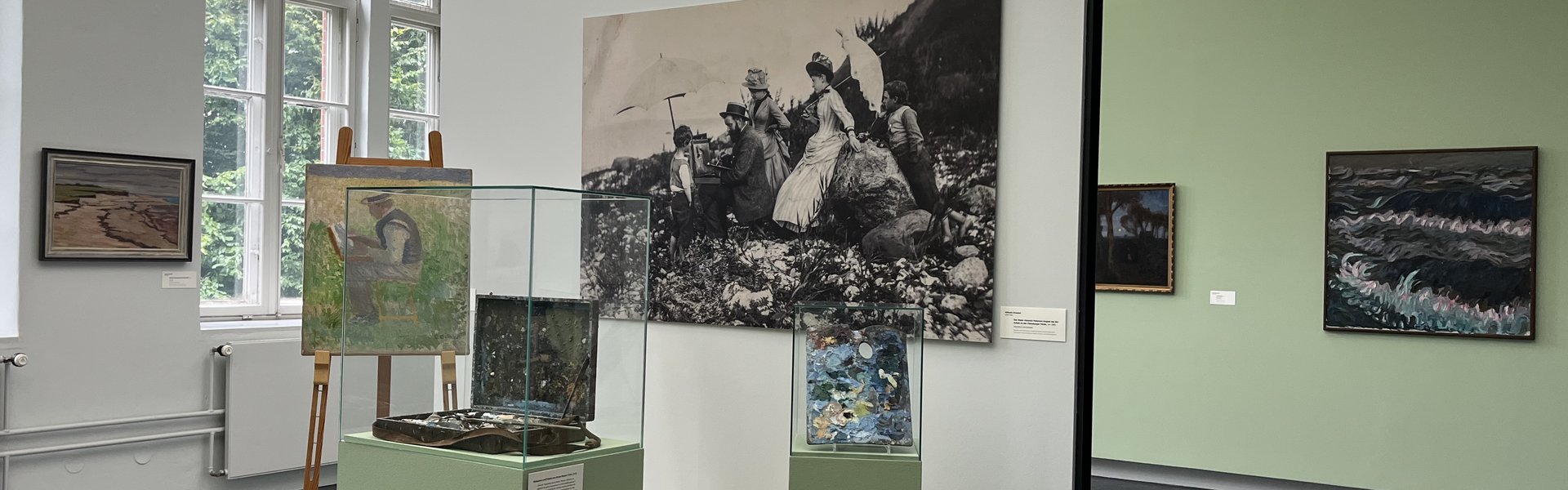 Eine bunte Fotografie. Ein Museumsraum. Im Hintergrund ist auf der linken Seite ein altes großes Fenster. Auf der rechten Seite hängen zwei Gemälde an der Wand. Im Vordergrund ist ein kleines Podest mit Wand. An der Wand hängt eine große Schwarz-weiß-Fotografie eines Mannes der im Freien an einem Bild auf einer Staffelei arbeitet. Um ihn herum stehen Frauen und Kinder in Kleidern, die man um 1900 getragen hat. Auf dem Podest ist eine Staffelei. Darauf das Bild eines Mannes beim Malen. Daneben ist eine Vitrine mit einem alten Malkoffer. Ganz rechts ist eine kleinere Vitrine mit einer alten Malerpalette.  