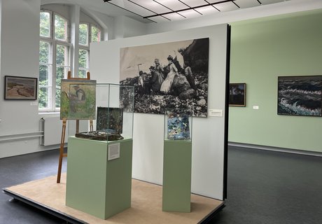 Eine bunte Fotografie. Ein Museumsraum. Im Hintergrund ist auf der linken Seite ein altes großes Fenster. Auf der rechten Seite hängen zwei Gemälde an der Wand. Im Vordergrund ist ein kleines Podest mit Wand. An der Wand hängt eine große Schwarz-weiß-Fotografie eines Mannes der im Freien an einem Bild auf einer Staffelei arbeitet. Um ihn herum stehen Frauen und Kinder in Kleidern, die man um 1900 getragen hat. Auf dem Podest ist eine Staffelei. Darauf das Bild eines Mannes beim Malen. Daneben ist eine Vitrine mit einem alten Malkoffer. Ganz rechts ist eine kleinere Vitrine mit einer alten Malerpalette.  