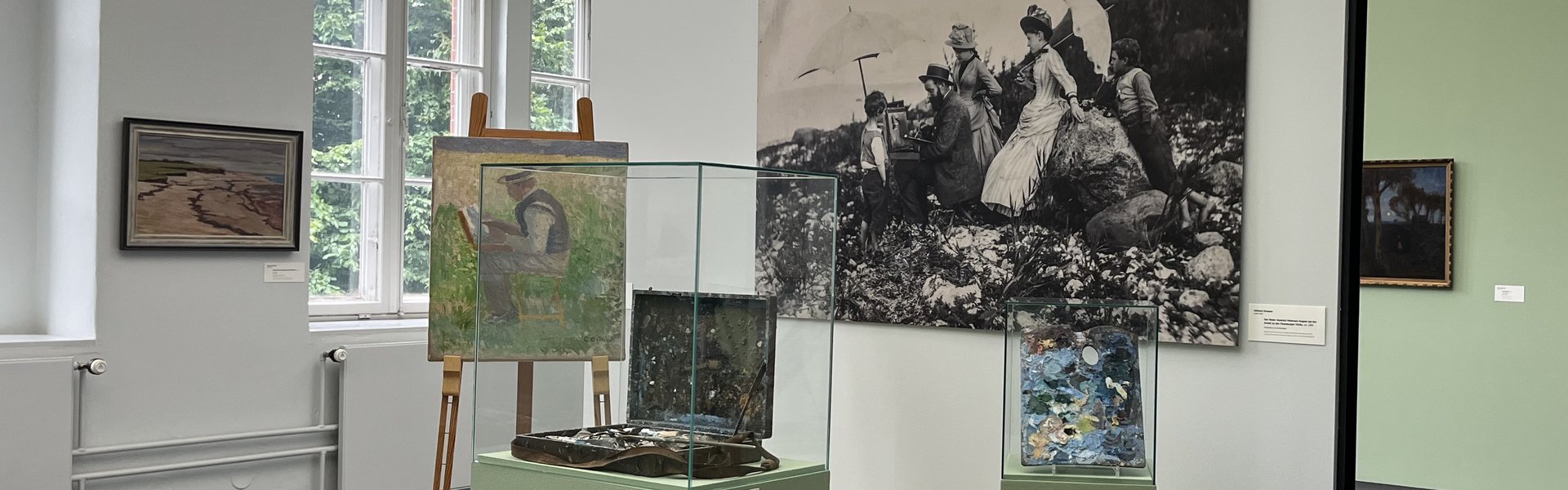 Eine bunte Fotografie. Ein Museumsraum. Im Hintergrund ist auf der linken Seite ein altes großes Fenster. Auf der rechten Seite hängen zwei Gemälde an der Wand. Im Vordergrund ist ein kleines Podest mit Wand. An der Wand hängt eine große Schwarz-weiß-Fotografie eines Mannes der im Freien an einem Bild auf einer Staffelei arbeitet. Um ihn herum stehen Frauen und Kinder in Kleidern, die man um 1900 getragen hat. Auf dem Podest ist eine Staffelei. Darauf das Bild eines Mannes beim Malen. Daneben ist eine Vitrine mit einem alten Malkoffer. Ganz rechts ist eine kleinere Vitrine mit einer alten Malerpalette.  