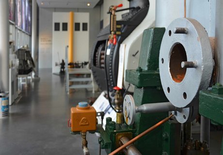 Blick durch die Technikhalle. Rechts und links an den Wänden stehen verschiedene Maschinen. Auch im Vordergrund ist eine große Maschine zu erkennen. Im Hintergrund ist die Brücke eines Schiffes angedeutet.