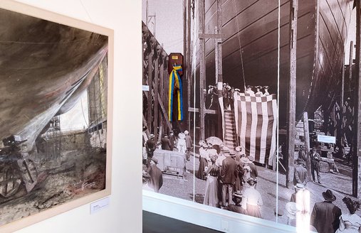 An einer Stell-Wand hängt ein modernes Gemälde. Man sieht: So sieht es in der Werft aus. Da wird ein Schiff gebaut. An der Wand rechts daneben hängt ein großes Foto. Es zeigt eine historische Schiffs-Taufe.