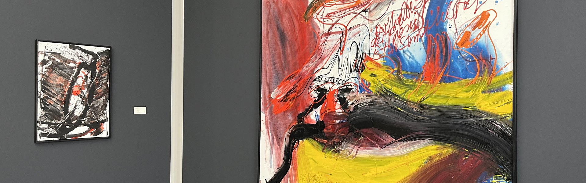 Auf der Fotografie sind zwei Gemälde vor einer grauen Wand zu sehen. Beide Bilder sind ungegenständlich. Das rechte zeigt auffällige rote, leuchtend gelbe und schwarze Farbverläufe. Das linke kräftige schwarze Linien und einige rote Farbfelder.