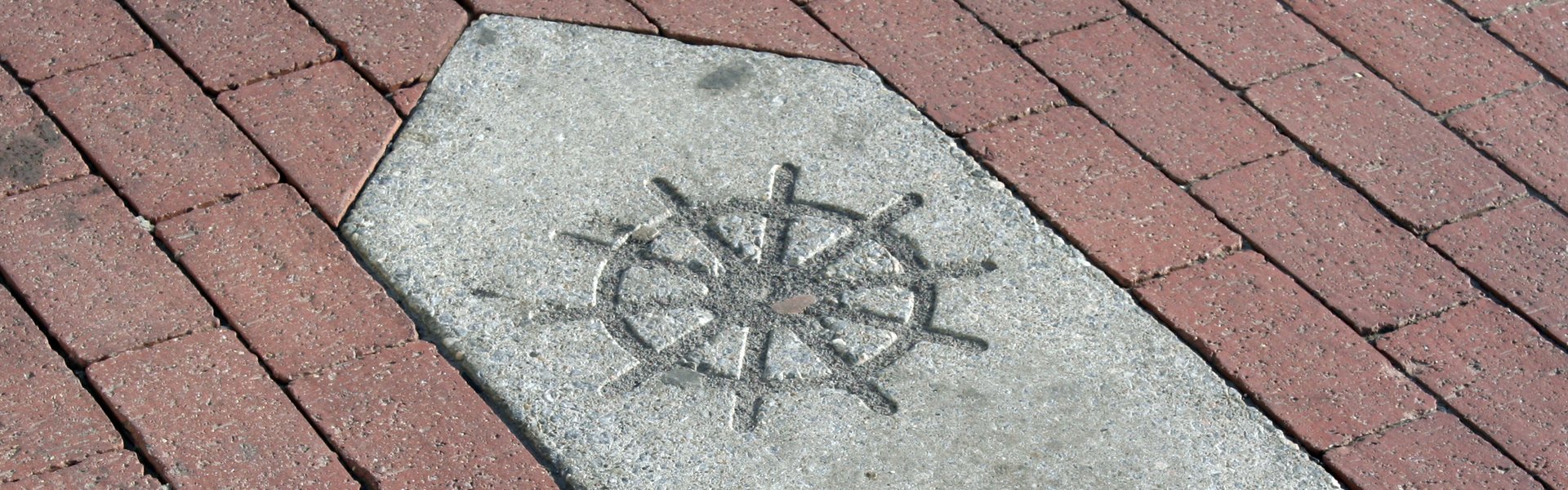 Man sieht einen Weg-Weiser aus Stein. Er ist für den KAPITÄNSWEG. Man findet ihn in der ganzen Stadt. Auf dem Weg-Weiser ist ein Pfeil und ein Steuer-Rad.