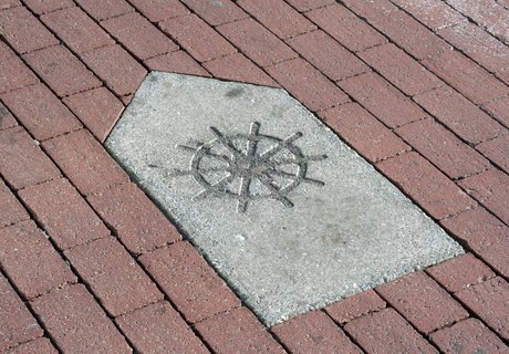 Man sieht einen Weg-Weiser aus Stein. Er ist für den KAPITÄNSWEG. Man findet ihn in der ganzen Stadt. Auf dem Weg-Weiser ist ein Pfeil und ein Steuer-Rad.