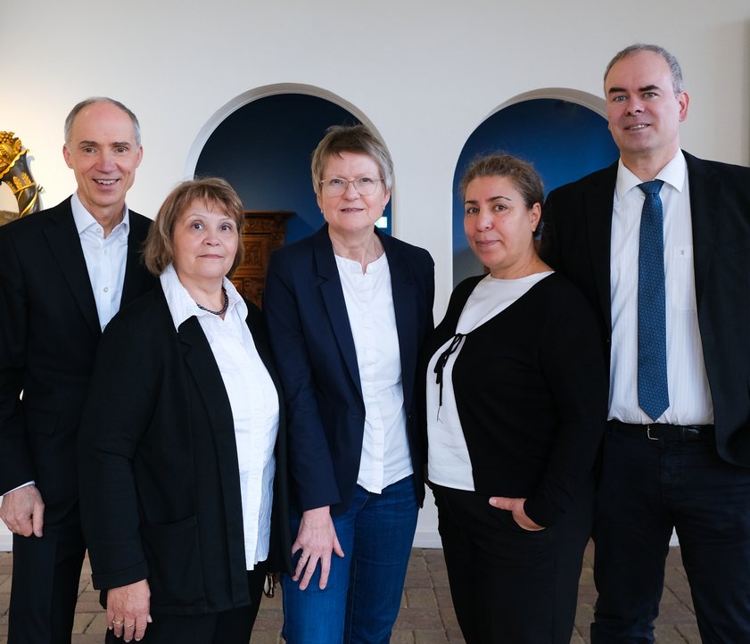 Fünf Personen in ordentlicher Kleidung stehen vor Kunstwerken in einem Museum. Drei Frauen sind von zwei Männern rechts und links umgeben.Sie lächeln freundlich in die Kamera. 