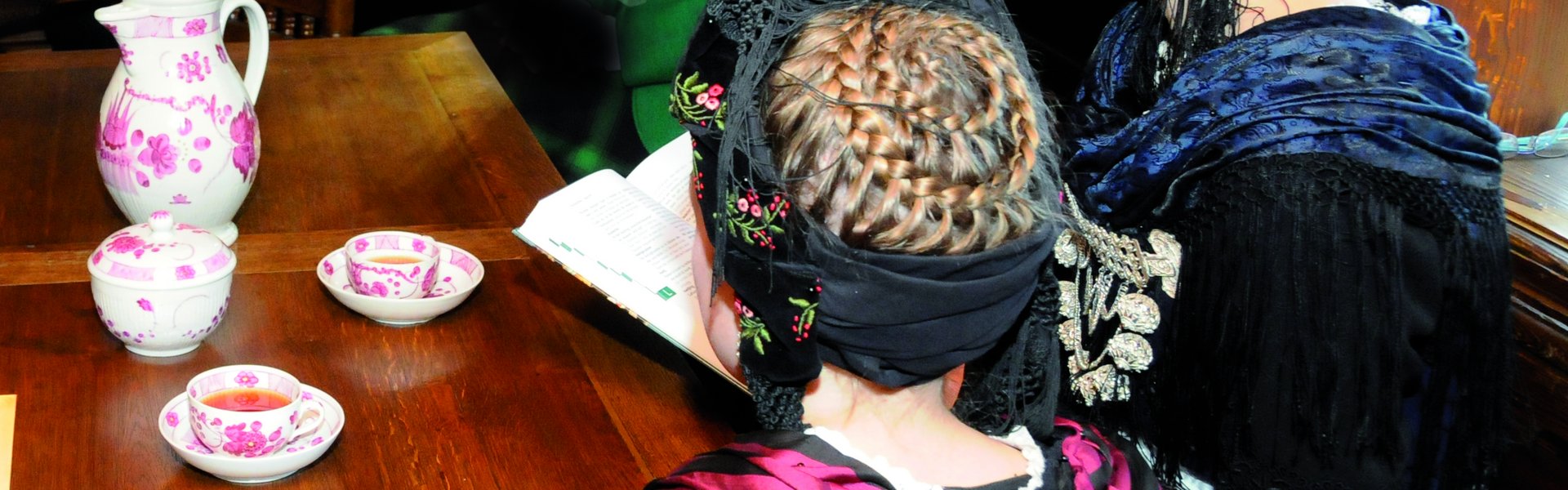 Es sind zwei Frauen in friesischer Tracht zu sehen, die Teetassen vor sich stehen haben.