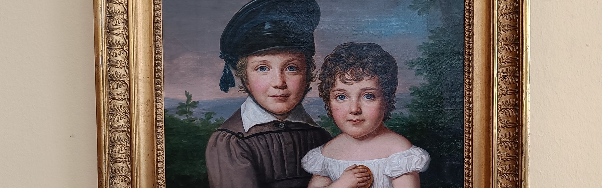 Auf der Fotografie sehen Sie eine helle Wand. An der Wand hängt ein Gemälde in einem verzierten Rahmen aus Messing. Das Gemälde zeigt zwei Kinder. Ein Junge trägt eine Mütze und ein hellbraunes Gewand. Das Mädchen trägt ein weißes Kleid. Auf ihrem Schoß steht ein Korb mit Äpfeln.