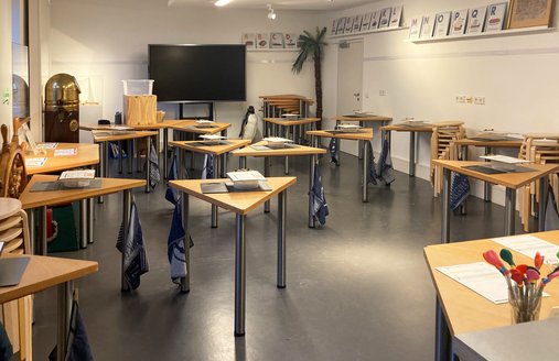 Eine bunte Fotografie. Darauf zu sehen ist ein Raum. Darin verteilt sind mehrere dreieckige Tische, auf denen Arbeitsmaterialien liegen. 