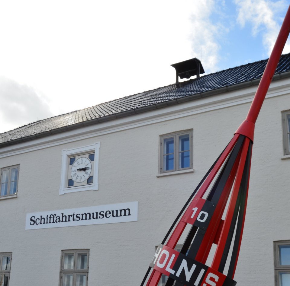 Vor dem Hauptgebäude des Flensburger Schifffahrtsmuseum steht ein markantes Seezeichen. Die Tonne ist rot und schwarz angemalt und trägt zudem die Aufschrift Holnis. Auch eine 10 ist auf die Seetonne gemalt. An der Fassade des Schifffahrtsmuseums hängt ein Schild mit der Aufschrift: Schiffahrtsmuseum. Darüber hängt eine Uhr. Die Fenster des Museums haben alle Sprossen.