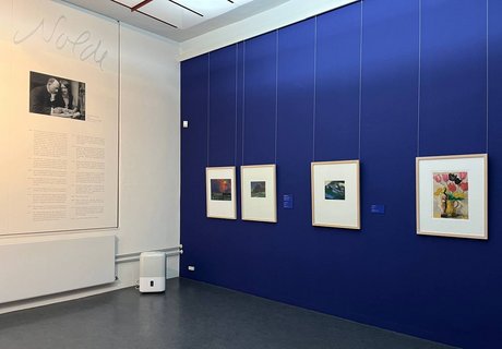 Auf dem Bild sieht man eine Ecke in einem Ausstellungsraum im Museumsberg Flensburg. Auf der linken Seite hängt ein Text, der von dem Künstler Emil Nolde handelt. Über dem Text ist ein schwarz-weißes Foto von einem Mann und einer Frau angebracht. Die Wand auf der rechten Seite ist dunkelblau gestrichen. Darauf hängen vier bunte Bilder mit braunen Holzrahmen.