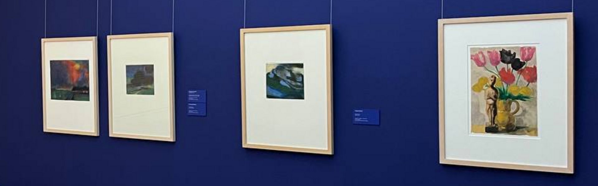 Eine bunte Fotografie. Darauf zu sehen ist die Zimmerecke eines Ausstellungsraums des Museumsberg Flensburgs. Auf der linken Seite ist ein Ausstellungstext zu sehen. Teil der Überschrift lautet „Nolde“. Über dem Text ist eine Schwarz-Weiß-Fotografie von einem Mann und einer Frau zu erkennen. Rechts davon ist die Wand in einem dunklen Blau gestrichen. An ihr hängen vier bunte Gemälde. Jedes Gemälde ist in einem hellbraunen Holzrahmen. 