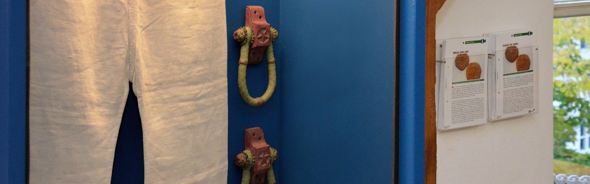 På farvebilledet ses et par sømandsbukser i en blå hængende vitrine. Ved siden af – ligeledes på væggen – hænger to gennemsigtige kortbeholdere. I disse er samlingskort sorteret. Disse består af et billede og tekst.