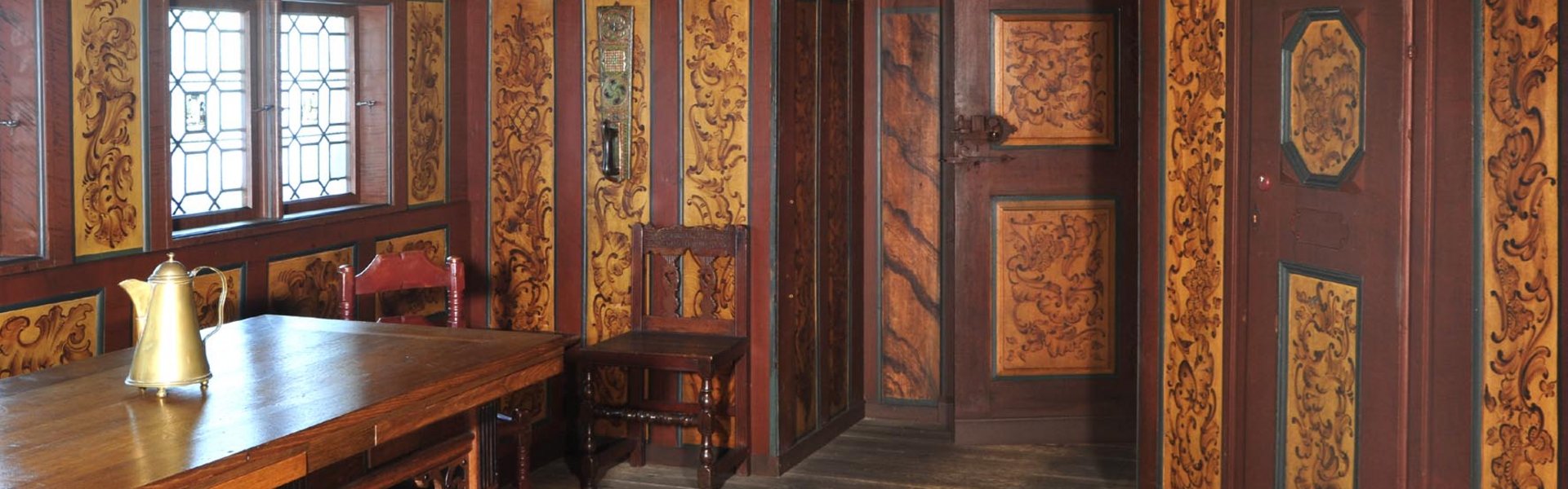 An den Wänden und der Decke des Raumes ist Holz mit Verzierungen angebracht. Ein Stuhl und ein Tisch stehen darin.