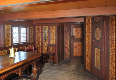 An den Wänden und der Decke des Raumes ist Holz mit Verzierungen angebracht. Ein Stuhl und ein Tisch stehen darin.