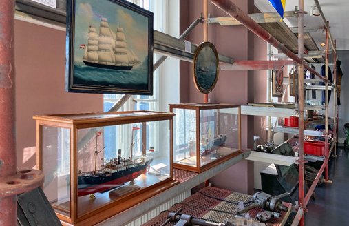 Auf dem Foto ist das lange Metall-Regal abgebildet. Man sieht auch zwei Schiffs-Bilder am Regal. Das sind die ersten Schiffe, die die FSG gebaut hat. Es sind Holz-Schiffe mit Segeln. Später baute die FSG auch Stahl-Schiffe. 