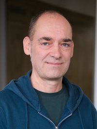 Portrait eines Mannes. Er trägt ein grünes T-Shirt und eine blaue Pullover-Jacke. Der Mann lächelt in die Kamera.