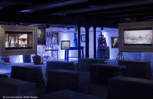 I forgrunden af billedet ses betonbænke. Disse fungerer som siddepladser under filmvisningen i romkælderen. Filmen vises på skærme. På dette billede kan man se to af de tre skærme, der hænger ned fra loftet.