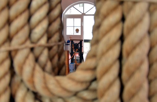 Vorne hängt ein dickes und langes Seil von der Decke. Dahinter kann man die Reep-Schlag-Maschine sehen. Man sieht auch die offene Holztür. Sie führt auf den Innen-Hof vom Flensburger Schifffahrts-Museum.