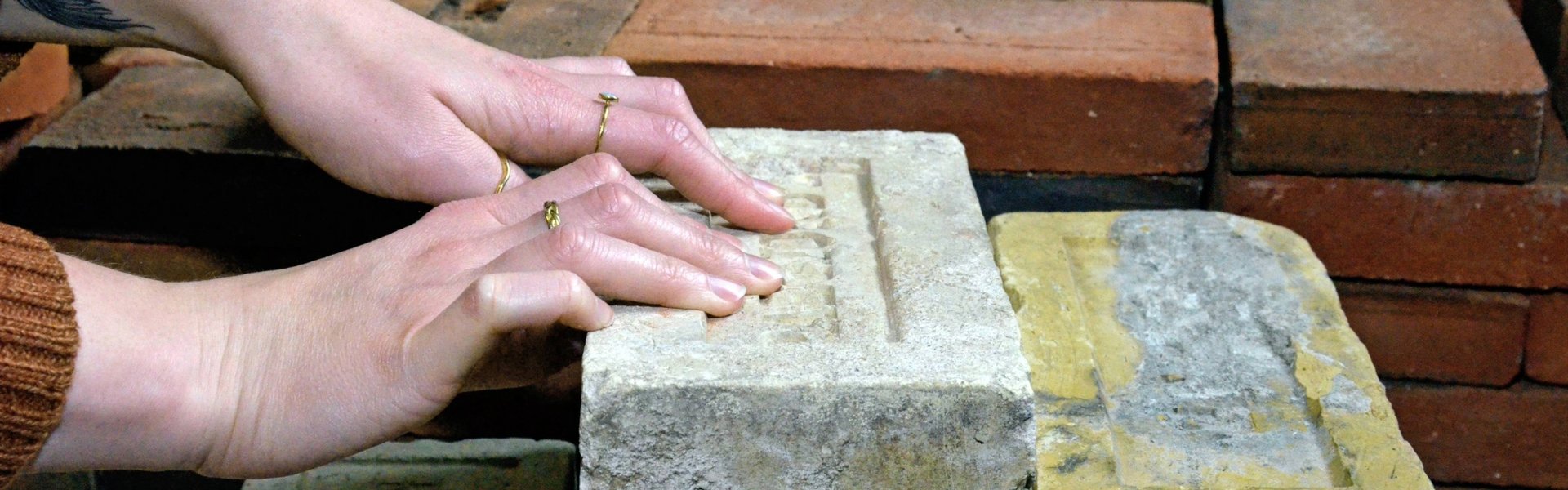 På farvefotografiet ses mursten. De har forskellige størrelser og farver. Nogle er hvide, andre gule og andre røde. Murstenene er stablet oven på hinanden. Der er flere stakke. To hænder rører ved en af de større sten. Der er indgraveret et ord på stenen.
