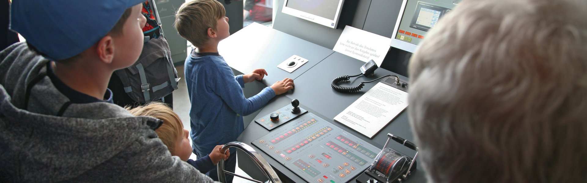 På farvefotoet ses fem personer: to voksne og tre børn. Børnene står lige ved skibssimulatoren. To af dem holder roret. De får hjælp af en hvidhåret mand.