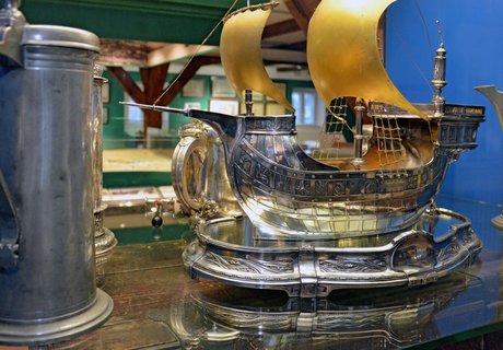 [Translate to Leichte Sprache:] Auf dem Farbfoto ist der Silberschatz des Schiffergelages zu sehen. Er befindet sich in einer Vitrine. Zu sehen ist ein aus Silber geformtes Segelschiff sowie einige Trinkgefäße. Hinter der Vitrine kann schemenhaft das Stadtmodell erahnt werden.