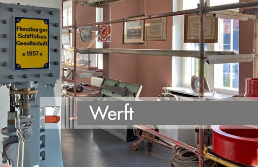 Links steht eine Maschine mit einem gelben Schild. Auf diesem steht: Flensburger Schiffsbau Gesellschaft 1957. Links daneben steht ein langes Metall-Regal. Da hängen Bilder. Außerdem liegen hier Werkzeuge.