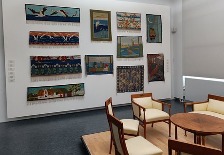 Auf der bunten Fotografie sieht man Teile eines Ausstellungsraums. Rechts im Vordergrund sind Möbel im Jugendstil zu sehen: Drei Stühle aus Holz mit weißen Polstern, eine Bank aus Holz mit weißen Polstern und ein kleiner runder Tisch in der Mitte. Hans Christiansen hat die Möbel entworfen. Hinten ist auf der rechten Seite ein Teil eines großen Fensters. Es ist mit einem weißen Rollo verhängt. Links ist eine weiße Wand. An der Wand hängen viele bunte Teppiche. Die Teppiche haben verschiedene Muster und sind handgewebte Scherrebek-Teppiche. Scherrebek ist ein Ort, der für Weberei berühmt ist.