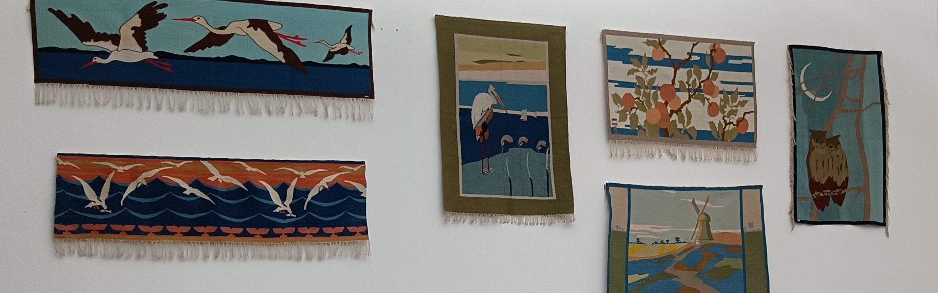 Auf der bunten Fotografie sieht man Teile eines Ausstellungsraums. Rechts im Vordergrund sind Möbel im Jugendstil zu sehen: Drei Stühle aus Holz mit weißen Polstern, eine Bank aus Holz mit weißen Polstern und ein kleiner runder Tisch in der Mitte. Hans Christiansen hat die Möbel entworfen. Hinten ist auf der rechten Seite ein Teil eines großen Fensters. Es ist mit einem weißen Rollo verhängt. Links ist eine weiße Wand. An der Wand hängen viele bunte Teppiche. Die Teppiche haben verschiedene Muster und sind handgewebte Scherrebek-Teppiche. Scherrebek ist ein Ort, der für Weberei berühmt ist.