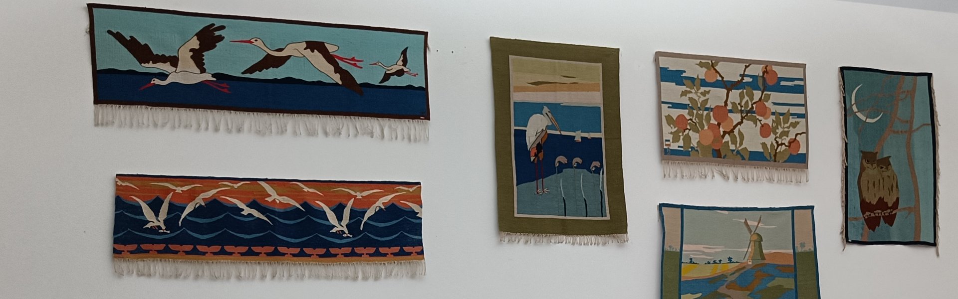 Eine bunte Fotografie. Darauf sind Teile eines Ausstellungsraums zu sehen. Rechts im Vordergrund sieht man Teile einer Möbelgarnitur. Drei Stühle aus Holz mit weißen Polstern, eine Bank aus Holz mit weißen Polstern und in der Mitte ein runder kleiner Tisch. Alles im Jugendstil. Die Garnitur ist von Hans Christiansen entworfen. Im Hintergrund sieht man auf der rechten Seite Teile eines großen Fensters, das mit einem weißen Rollo verhangen ist. Auf der linken Seite ist eine weiße Wand, an der viele bunte Teppiche mit unterschiedlichen Motiven hängen. Die Teppiche sind handgewebte Scherrebek Teppiche. 