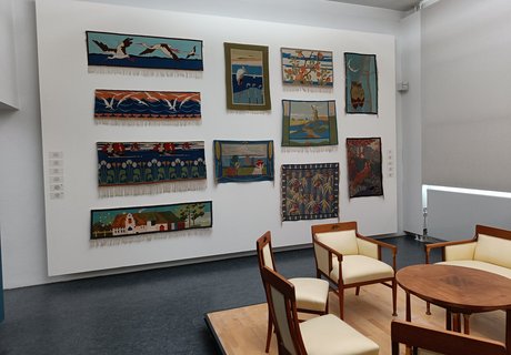 Eine bunte Fotografie. Darauf sind Teile eines Ausstellungsraums zu sehen. Rechts im Vordergrund sieht man Teile einer Möbelgarnitur. Drei Stühle aus Holz mit weißen Polstern, eine Bank aus Holz mit weißen Polstern und in der Mitte ein runder kleiner Tisch. Alles im Jugendstil. Die Garnitur ist von Hans Christiansen entworfen. Im Hintergrund sieht man auf der rechten Seite Teile eines großen Fensters, das mit einem weißen Rollo verhangen ist. Auf der linken Seite ist eine weiße Wand, an der viele bunte Teppiche mit unterschiedlichen Motiven hängen. Die Teppiche sind handgewebte Scherrebek Teppiche. 