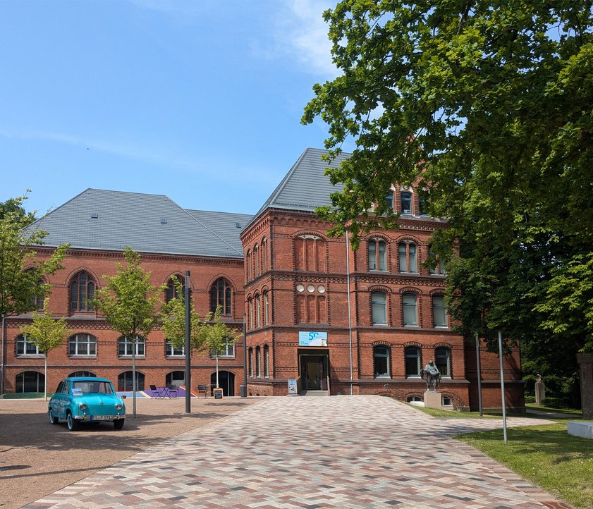 Das rote Gebäude ist das Hans-Christiansen-Haus. Davor stehen ein blaues Auto und eine Skulptur. Ein Weg führt zum Eingang. Daneben sind Bäume.