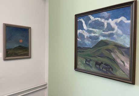 Das Foto zeigt zwei Gemälde von Käte Lassen. Sie hängen in einem Ausstellungsraum vor einer mintgrünen Wand. Auf dem rechten Gemälde ist eine hügelige Landschaft zu sehen. In der Bildmitte befindet sich ein orangefarbener, leuchtender Mond. Auf dem linken Gemälde ist ebenfalls eine hügelige Landschaft zu sehen. Über dem Horizont ist der blaue Himmel mit weißen Wolken dargestellt. In der Landschaft im Bildvordergrund befinden sich vier Schafe.  
