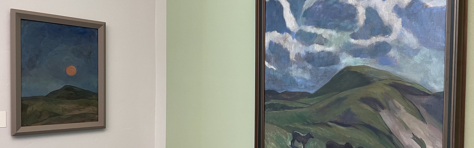 Das Foto zeigt zwei Gemälde von Käte Lassen. Sie hängen in einem Raum mit einer mintgrünen Wand. Auf dem rechten Gemälde sieht man eine hügelige Landschaft. In der Mitte des Bildes leuchtet ein orangefarbener Mond. Auf dem linken Gemälde ist auch eine hügelige Landschaft zu sehen. Über dem Horizont ist der blaue Himmel mit weißen Wolken. Vorne stehen vier Schafe.